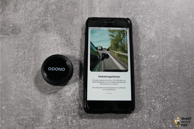 Der Ooono CO-DRIVER NO1 liegt neben dem Smartphone und wird gedrückt, um eine Verkehrswarnung zu übermitteln.